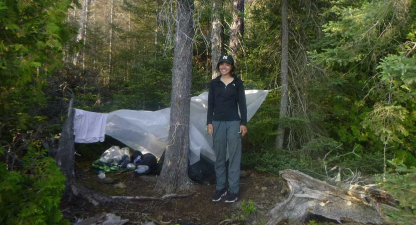a person stands beside their tarp shelter in a wooded area on an outward bound trip